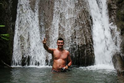 Portrait of man in river gesturing peace sign against waterfall