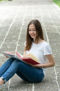 Smiling teenage girl writing in paper with book while sitting on footpath