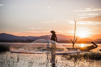 Man standing on boat by fishing net in lake against sky during sunset