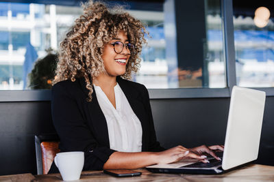 Smiling hispanic afro business woman in cafe working on laptop and mobile phone. tech and lifestyle