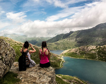 Rear view of women sitting on rock formation while looking at lake