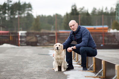 Portrait of man with golden retriever sitting on small fence during winter