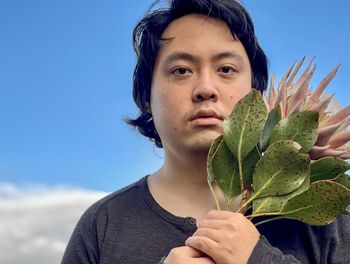 Close-up of young asian man holding pink king protea flower against blue sky with cloud