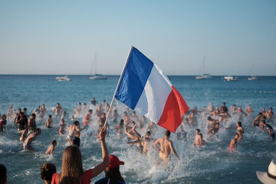 Woman holding french flag with people in sea against clear sky during sunset