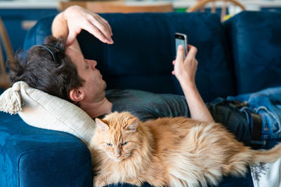 Adult man with smartphone and red furry cat spending a lazy afternoon on a blue couch. cozy home