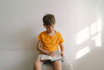 A cute boy wearing an orange t-shirt is sitting on a soft ottoman reading a book