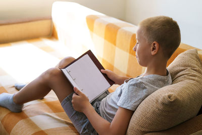 Caucasian school-age boy reading a book while sitting on the couch at home.