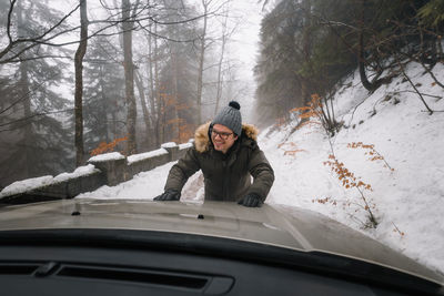 Man pushing car on snow covered road in foggy forest