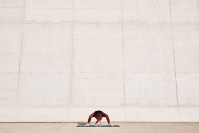 Unrecognizable fit female athlete in sportswear doing wide legged forward bend yoga pose on sports mat while training alone on street against concrete wall in sunny day