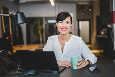 Portrait of confident businesswoman holding coffee mug while sitting at desk in office