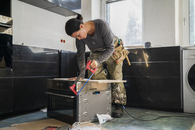 Female carpenter cutting plank on microwave in kitchen at apartment