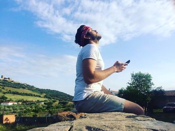 Side view of man holding mobile phone while relaxing on rock against sky