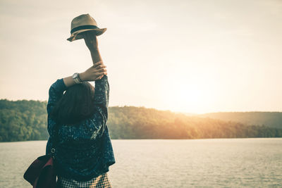 Rear view of woman holding hat at lakeshore against clear sky during sunset