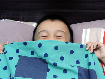 High angle portrait of boy covering mouth with turquoise sheet on bed