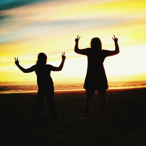 Rear view of silhouette mother with daughter showing peace sign at beach during sunset