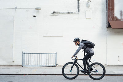 Side view of contemporary male cyclist in black helmet and eyeglasses surfing on mobile phone on white wall background