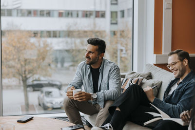 Smiling mature businessman with coffee cup sitting by male colleague during conference meeting