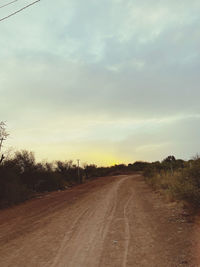 Dirt road along trees on field against sky