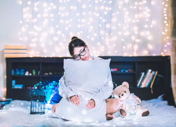 Young woman embracing pillow while sitting amidst christmas lights on bed