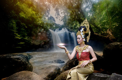 Beautiful young woman wearing crown and traditional clothing while sitting against waterfall in forest