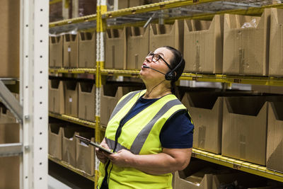 Mature female worker with digital tablet looking up while talking on headset against rack at distribution warehouse