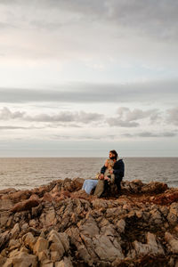 Bearded man embracing blond girlfriend in outerwear and looking away while sitting on stony coast of rippling sea on cloudy day in aviles, spain