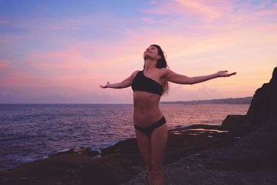 Woman with arms outstretched while standing at beach against sky during sunset