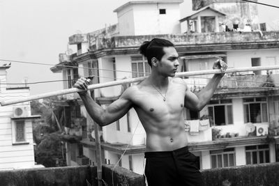 Shirtless young man carrying bamboo while standing against building in city
