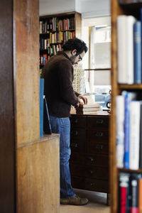 Man reading book while standing seen from shelves