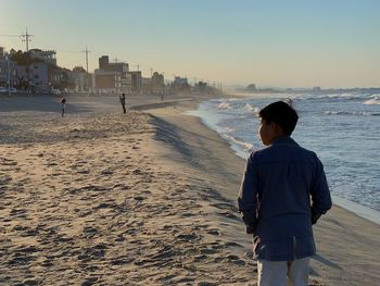 Rear view of boy standing at beach against clear sky during sunset