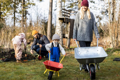 Smiling woman and son wheeling wheelbarrows while family gardening in background at back yard