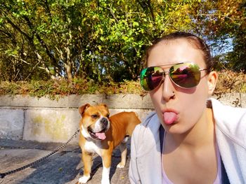 Portrait of woman wearing sunglasses and sitting out tongue with dog against retaining wall