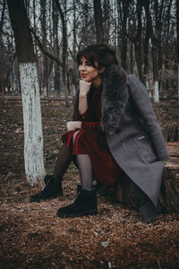 Full length of woman wearing fur coat sitting on tree stump in forest