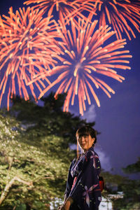 Low angle view of young woman standing against firework display at dusk