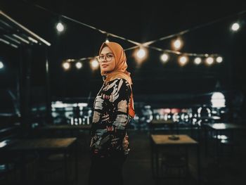 Portrait of woman in headscarf standing against illuminated lights at night