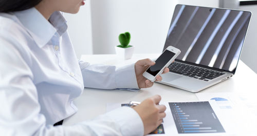 Midsection of businesswoman using smart phone sitting by laptop at office