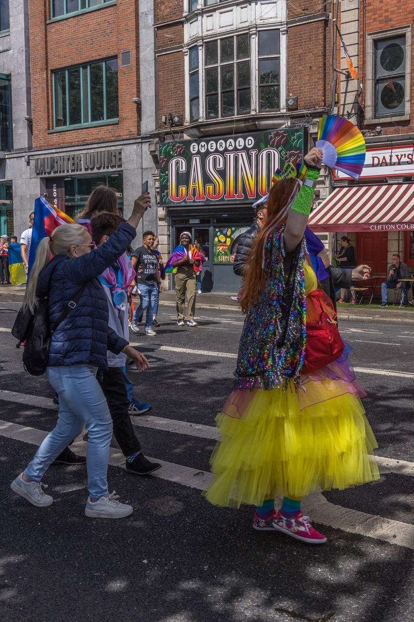 city, architecture, building exterior, street, group of people, full length, built structure, carnival, adult, women, event, celebration, men, arts culture and entertainment, tradition, festival, clothing, costume, performance, dancing, city life, crowd, lifestyles, leisure activity, traditional clothing, day, city street, multi colored, large group of people, outdoors