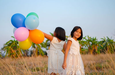 Portrait of girl standing with sister holding colorful balloons while standing on grassy field against clear sky