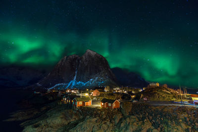 Illuminated houses by mountain against aurora borealis in sky at night