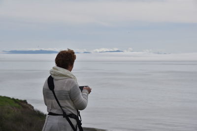 Rear view of woman photographing fog through smart phone against sky