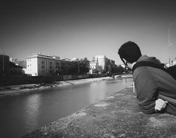 Rear view of man sitting on riverbank against clear sky