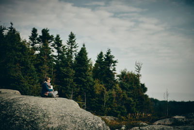 Man sitting on rock by trees against sky