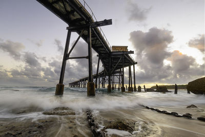 Low angle view of pier over beach against cloudy sky at sunset