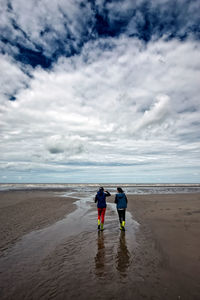 Rear view of friends walking at beach against cloudy sky