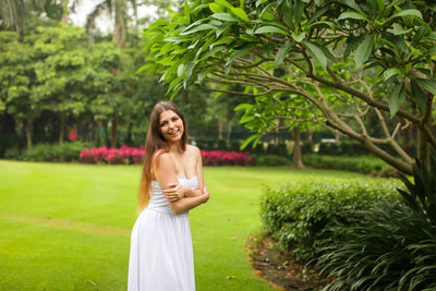 Portrait of smiling mid adult woman wearing off shoulder dress standing on grassy field against trees in park