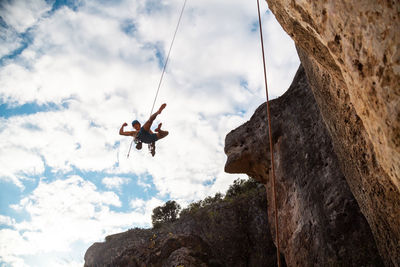 Low angle view of man hanging from safety harness by rock formation against sky