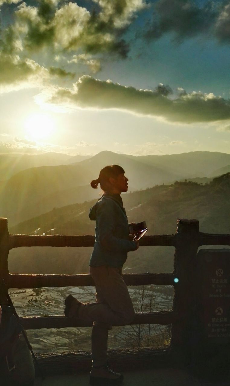 BOY STANDING ON MOUNTAIN AGAINST SKY AT SUNSET