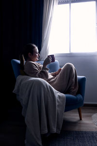 Woman drinking hot drink while sitting on the couch at home and staring the window