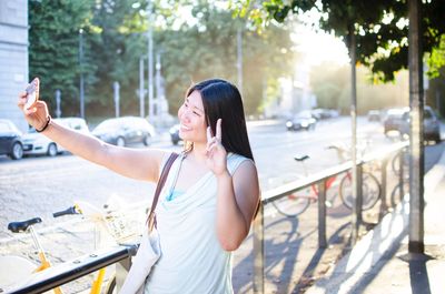 Woman gesturing while taking selfie through mobile phone on sidewalk