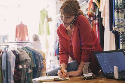 Woman writing order in diary while talking on smart phone at thrift store